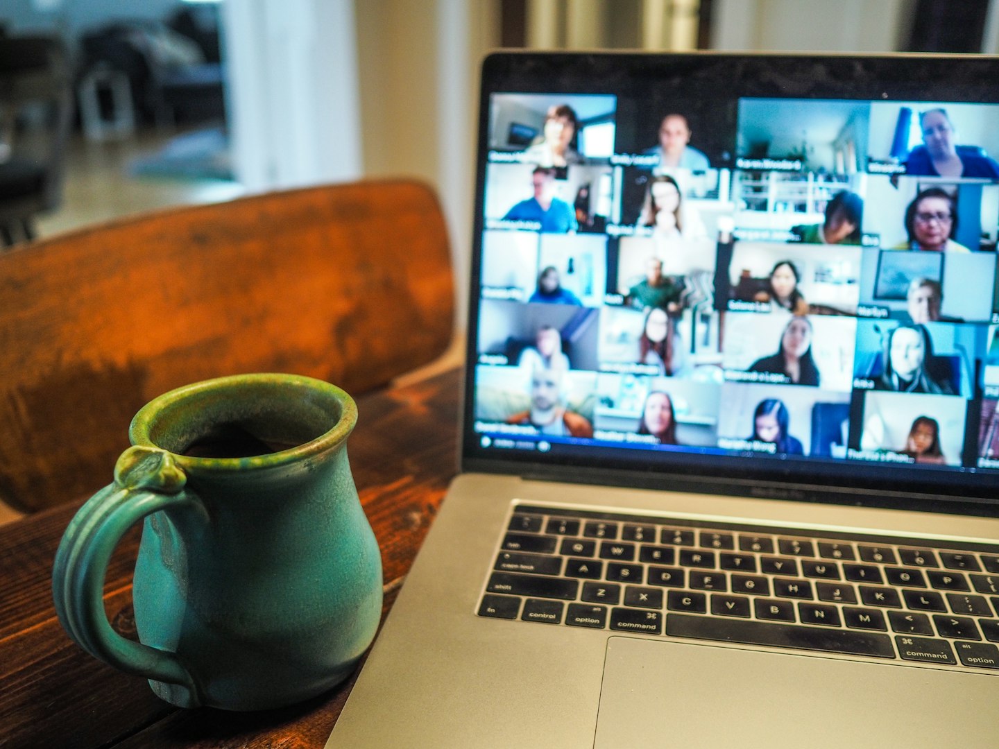 Ett hemmakontor med ett träbord i mörkt trä med en grön keramikmugg och en bärbar dator som visar ett videomöte på en föreläsning om ergonomi. PÅ bilden syns många deltagare som har egba skärmar.