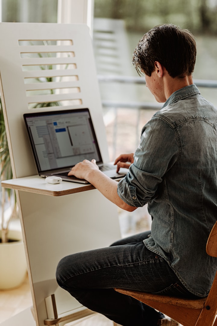 Man i gråblå skjorta sitter vid en bärbar dator i en upprätt ergonomisk position med hjälp av en ergonomisk utformat hjälpmedel